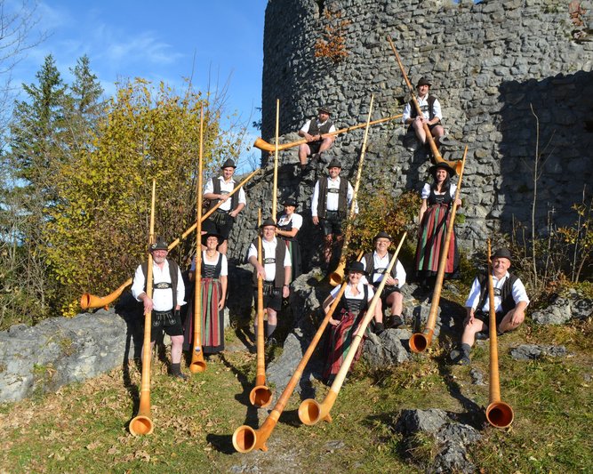 Zehn Personen in traditioneller Tracht stehen und sitzen um eine alte Steinmauer. Sie halten große Alphörner. Die Männer tragen schwarze Hüte und weiße Hemden, die Frauen bunte Röcke.