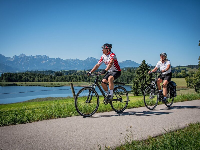 Zwei Radfahrer auf einem asphaltierten Weg am Illasbergsee, umgeben von grünen Wiesen und Bergen im Hintergrund.